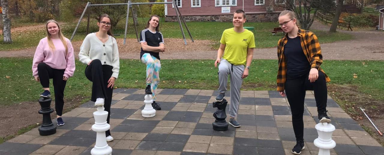 Viisi nuorta seisoo isolla shakkilaudalla, kullakin toinen jalka ison shakkinappulan päällä.